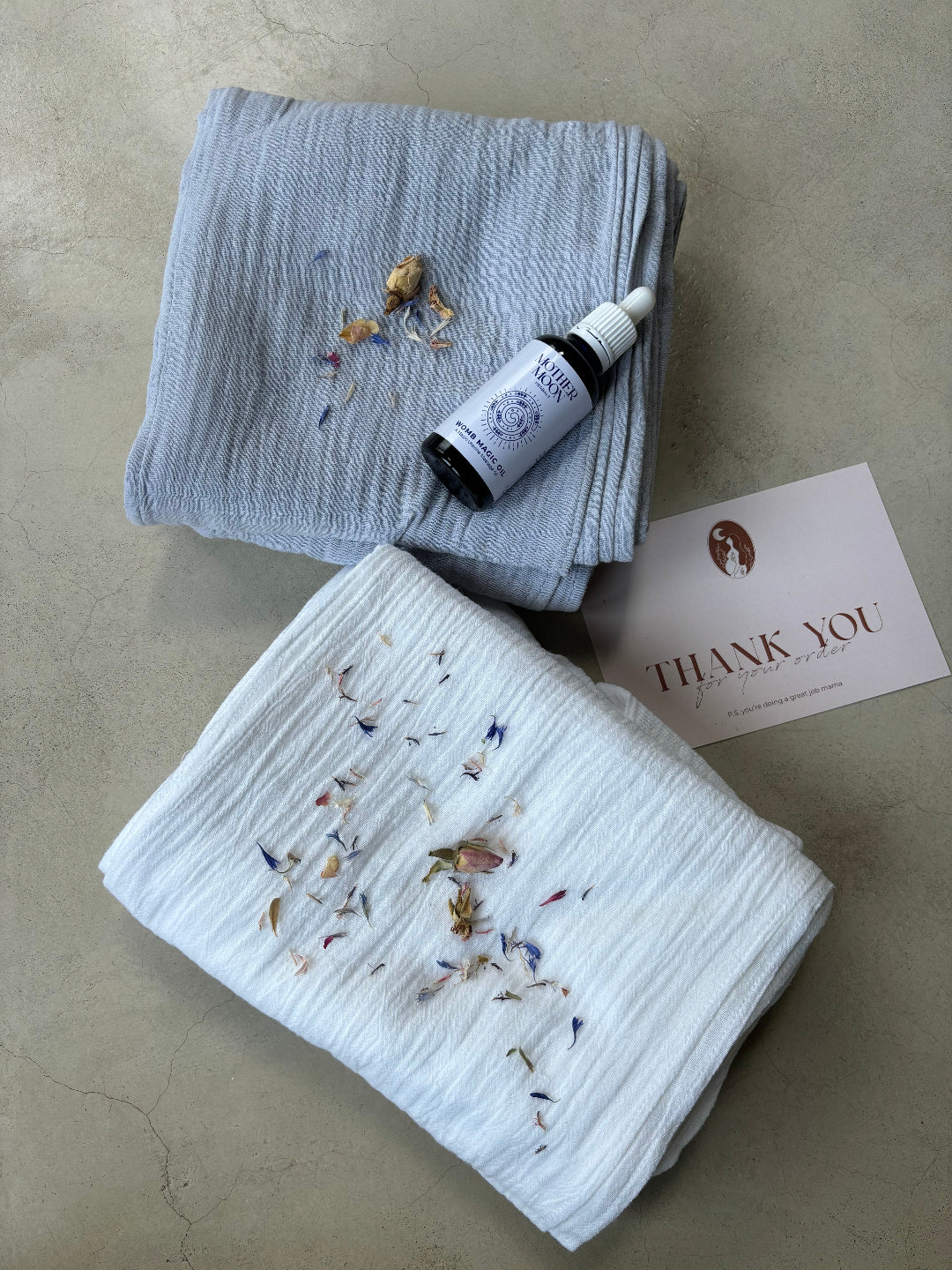 Two folded towels with dried flowers and Womb Magic Oil on a beige surface.
